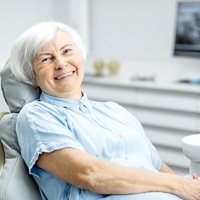 An older woman lying back in a dentist’s chair and smiling after having her dental implants put into place