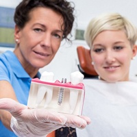 A dentist holding a cross-section of two natural teeth and a dental implant while the female patient looks and smiles