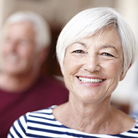 An older woman smiling while her husband smiles in the background
